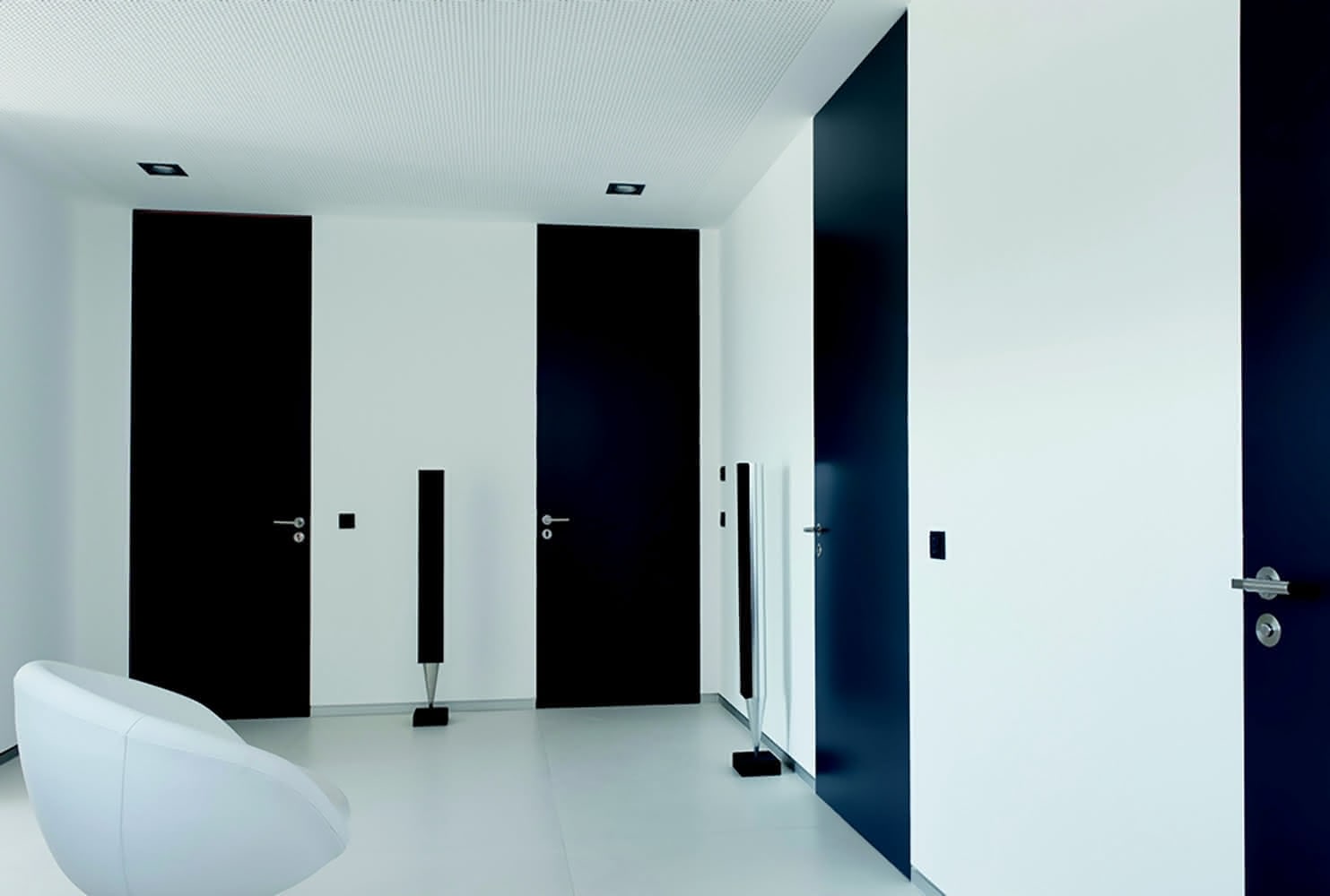 The doors in the image are sleek, floor-to-ceiling black doors that create a striking contrast against the white walls, giving the minimalist space an elegant, modern ambiance.